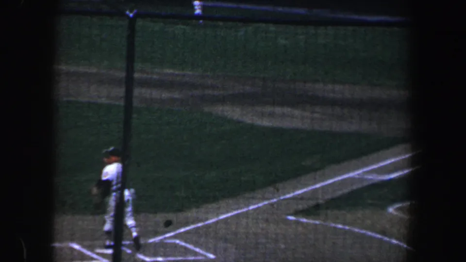 A uniformed player stands near the chalked batter's box during a 1960 Baltimore baseball game, seen from an elevated spectator angle.
