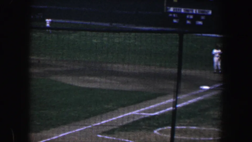 Wide 1960 Baltimore baseball view through stadium netting, showing chalk lines, a base path, a distant fielder, and a small scoreboard panel.