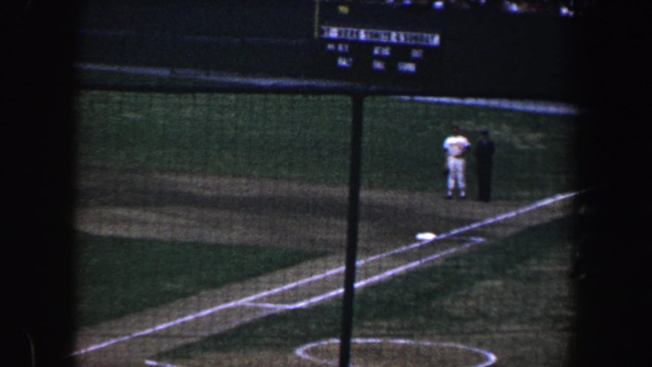 Night baseball field viewed through netting, with two figures near a baseline.