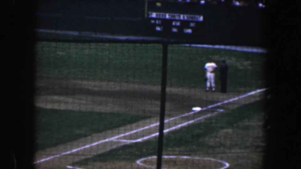 1960 Baltimore home-movie view of a night baseball field through protective netting, with two distant figures near the baseline.