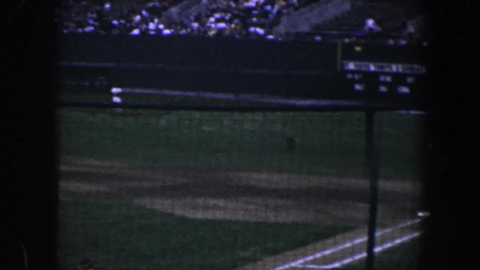 Night baseball infield in Baltimore, seen through netting with a distant player and scoreboard area in the background.