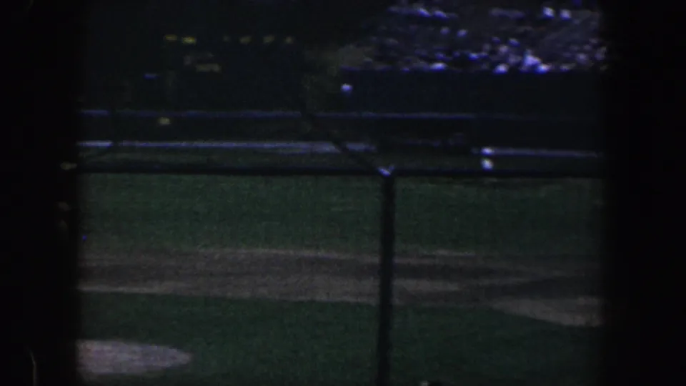 Grainy 8mm view of a dim nighttime baseball field, with diamond lines, netting, and distant spectator seating visible.