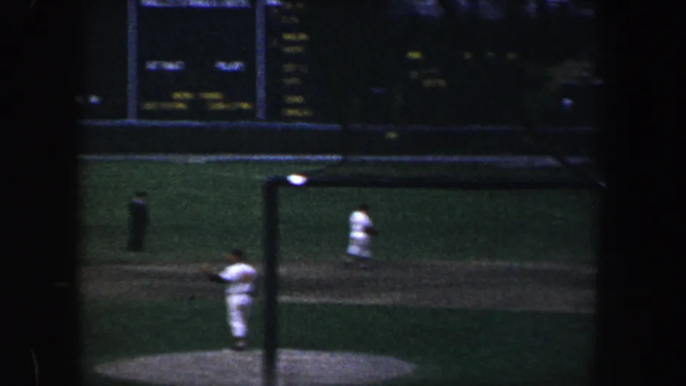 1960 Baltimore night baseball view through netting, with a pitcher on the mound and other distant players on the field.
