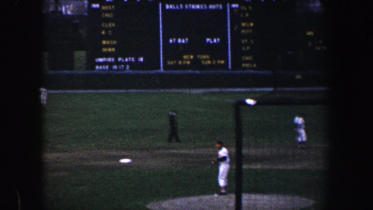 1960 Baltimore baseball footage showing a player on the mound, fielders in the distance, and an illuminated stadium scoreboard.