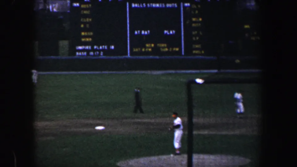 1960 Baltimore baseball footage showing a player on the mound, fielders in the distance, and an illuminated stadium scoreboard.