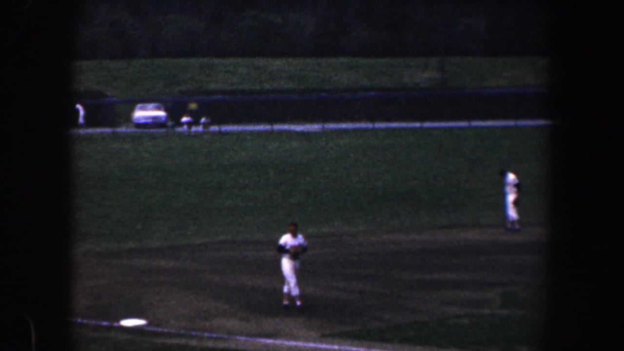 1960 Baltimore home-movie frame showing a youth baseball player on the infield, with a base, grass field, road, and parked car in the background.