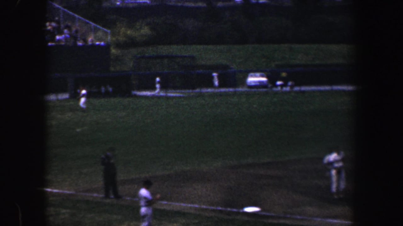 Baseball field seen from a distance with players, an umpire figure, and parked cars beyond the grass.