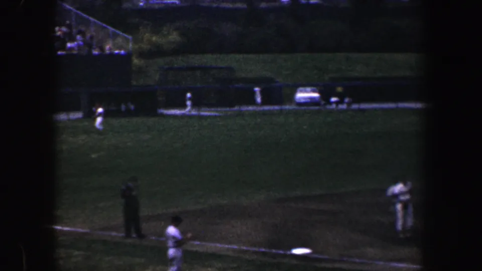 1960 Baltimore frame of a youth baseball game from a distant handheld viewpoint, showing players around the infield and cars along the far side.