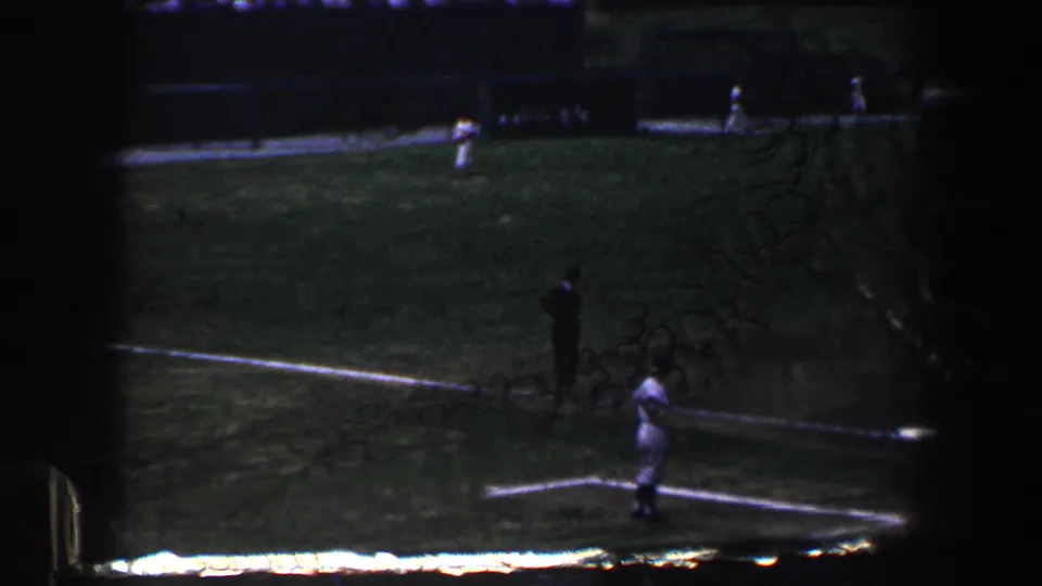 1960 Baltimore home-movie frame showing youth baseball action from above the field, with players near the baseline and a dark-clothed figure nearby.