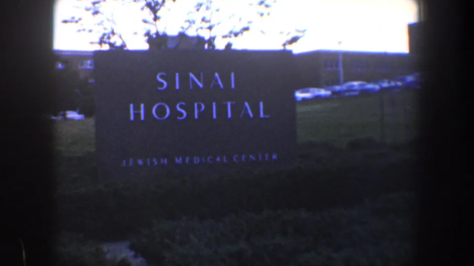1960 Baltimore frame showing a large Sinai Hospital Jewish Medical Center sign, with cars and landscaping visible around it.