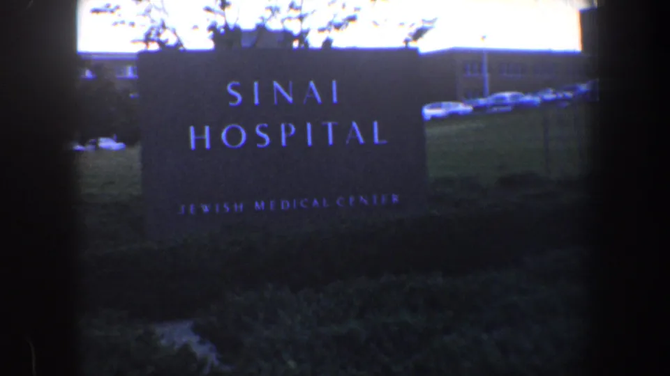 1960 Baltimore 8mm view of the Sinai Hospital sign on landscaped grounds, with hospital buildings and cars behind.