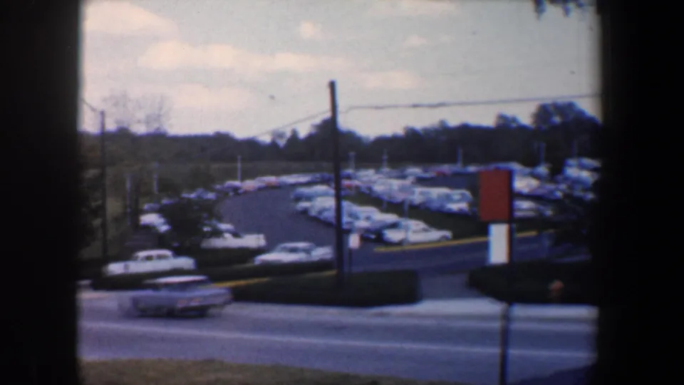 1960 Baltimore roadside view near Sinai Hospital, showing a large parking lot, utility poles, and a light-colored sedan in motion.