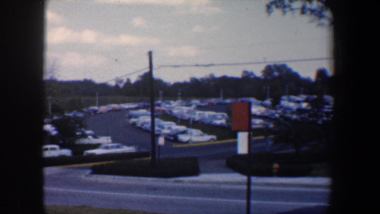 Roadway beside a parking lot filled with cars, with poles, trees, and a red sign visible.
