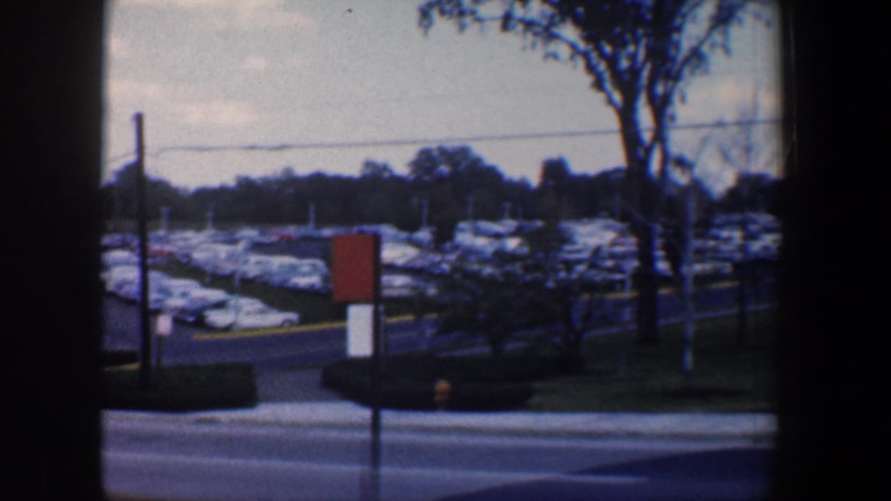 Parking lot with many cars seen beyond a road, framed by poles, trees, and dark film borders.