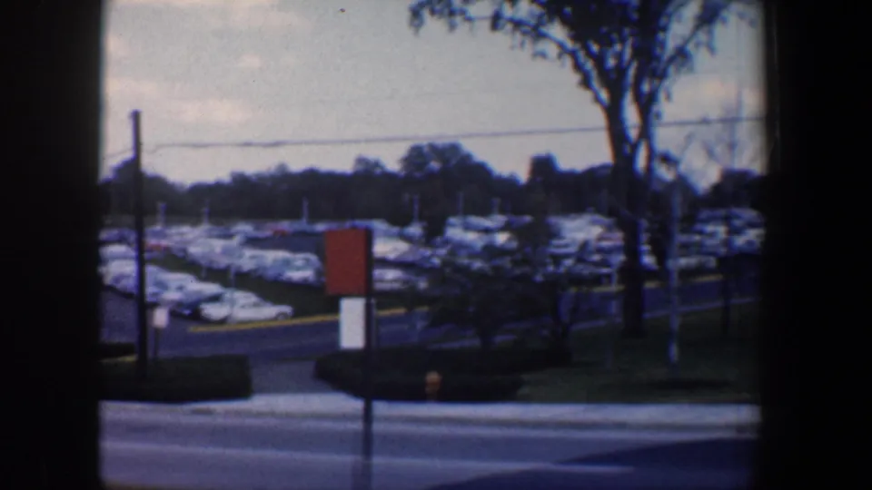 Baltimore parking-lot view during the rightward pan, showing rows of mid-century cars, a road, signpost, poles, and trees.