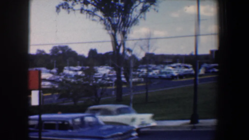 A light-colored mid-century car passes through the foreground while the Baltimore parking lot remains visible behind it.
