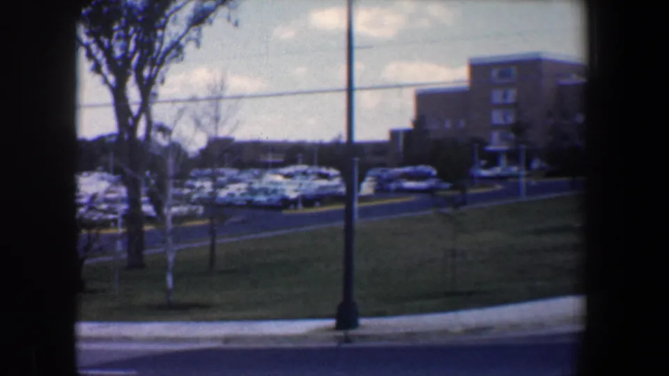 The pan reaches a wider parking-lot view with grass, trees, roadside poles, parked cars, and a low building in Baltimore.