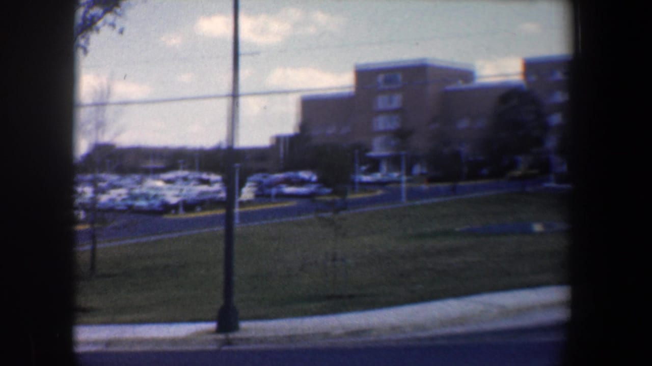 1960 Baltimore home-movie view of tan brick buildings, parked cars, lawn, sidewalk, and road seen through dark window-like borders.