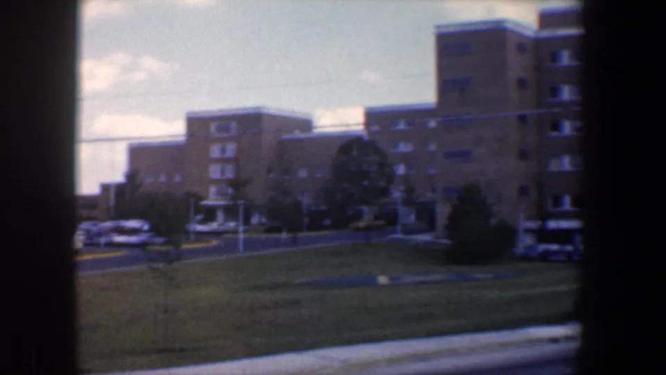 1960 Baltimore exterior view of a tan brick building complex with rows of windows, lawn, parked cars, and a road in front.