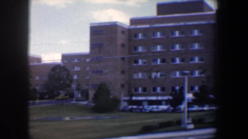 1960 Baltimore shot of a multi-story tan brick building with repeated windows, a front drive, parked cars, and lawn.