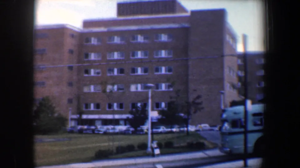 1960 Baltimore exterior with a tan brick building, parked cars, lawn, and a light blue bus partly visible at the right edge.