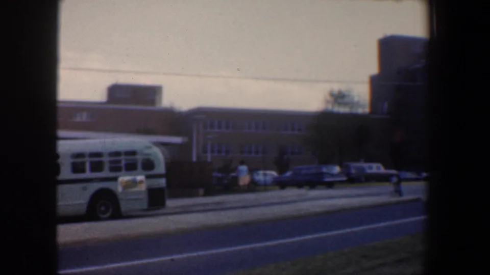 The bus exits the view, leaving a Baltimore street scene with brick institutional buildings and parked cars.