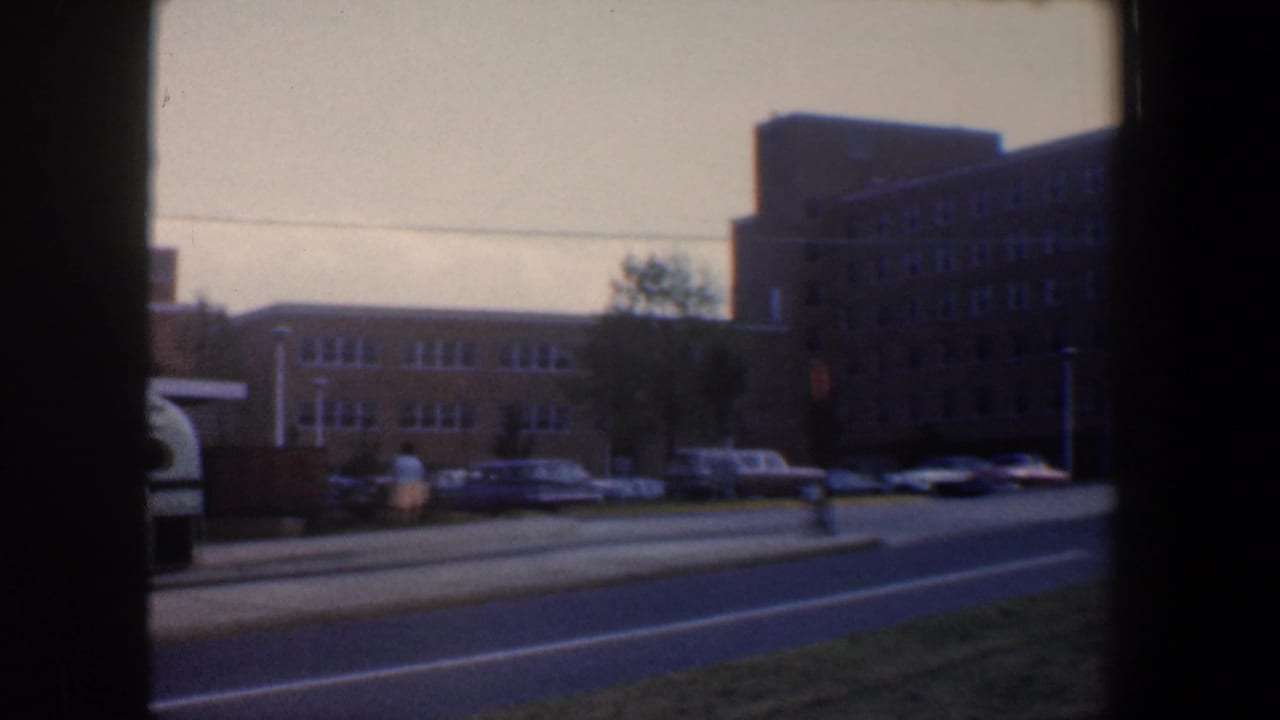 Tan brick building across a road with parked cars and grass in the foreground.