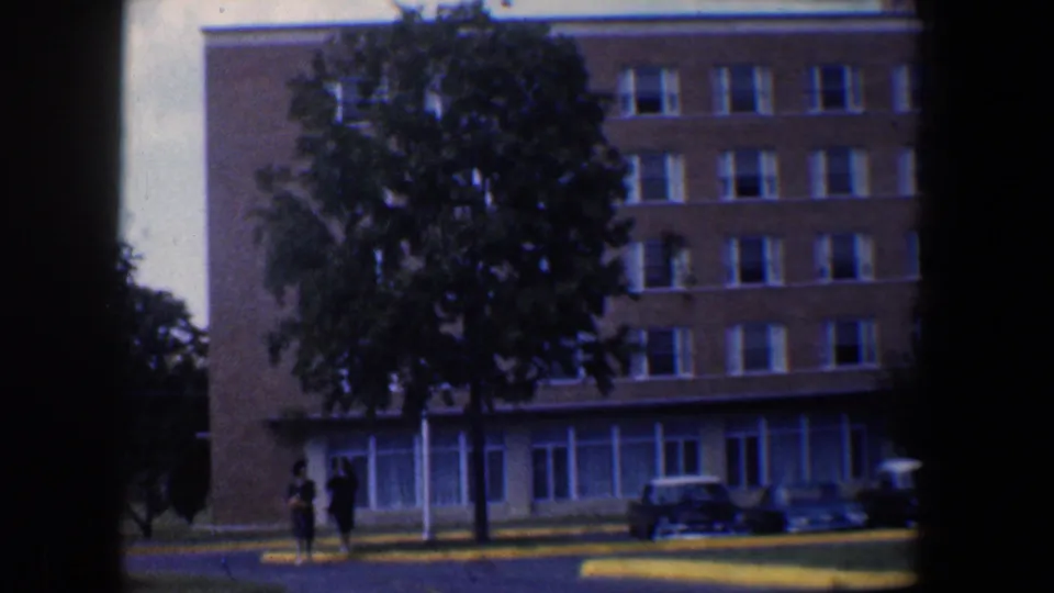 Closer 1960 Baltimore view of a tan brick building partly covered by a green tree, with distant pedestrians and parked cars.