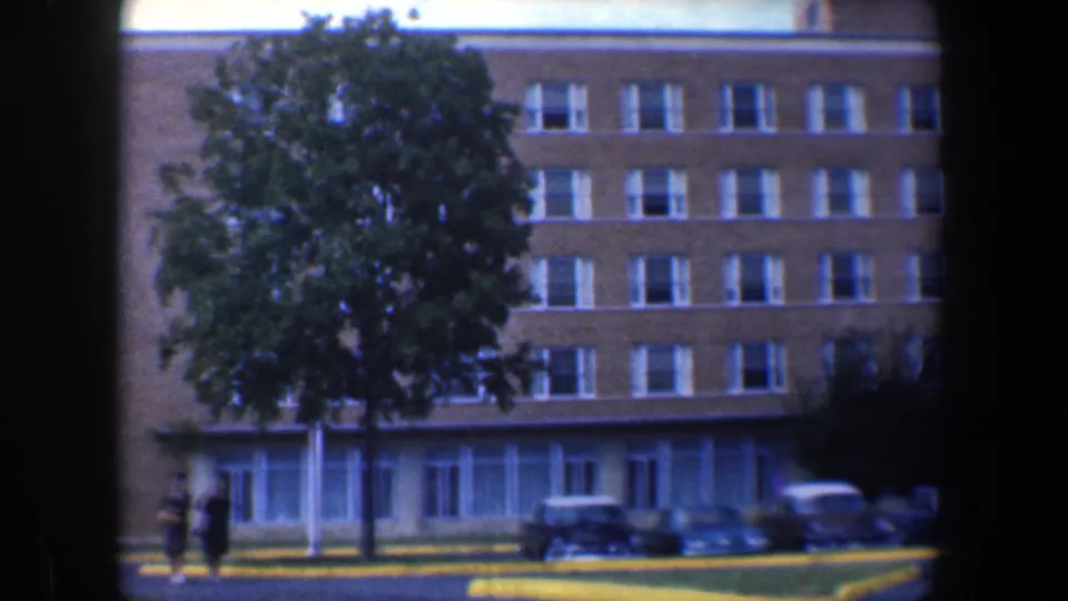 Baltimore building exterior with a prominent green tree, parked cars, sidewalk, and two small pedestrians in 1960 8mm footage.
