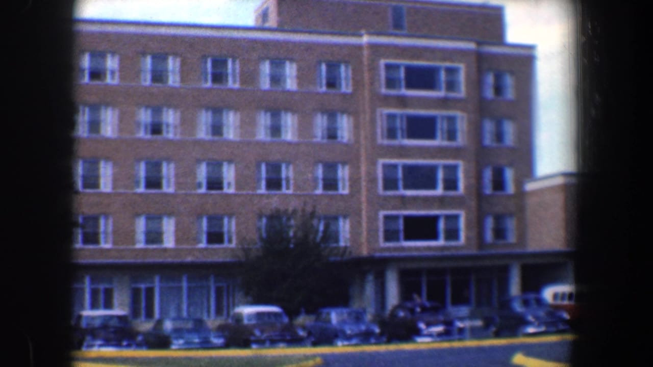 Wide view of the same brick building with parked cars, lawn, trees, and dark vertical frame edges.