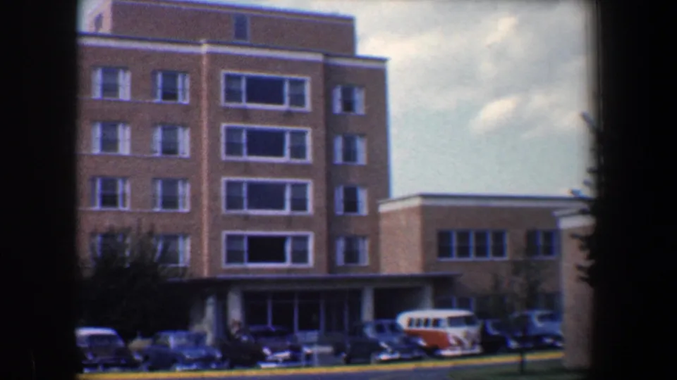 1960 Baltimore home-movie frame showing the building entrance, parked cars, and an orange-and-white van near the curb.