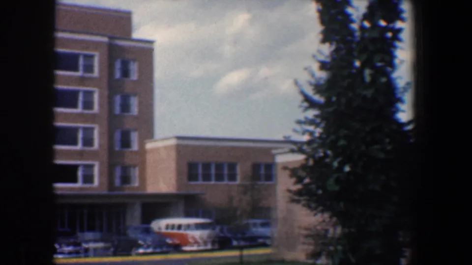 1960 Baltimore 8mm frame with the brick building corner, entrance canopy, parked cars, orange-and-white van, and tall evergreen tree.