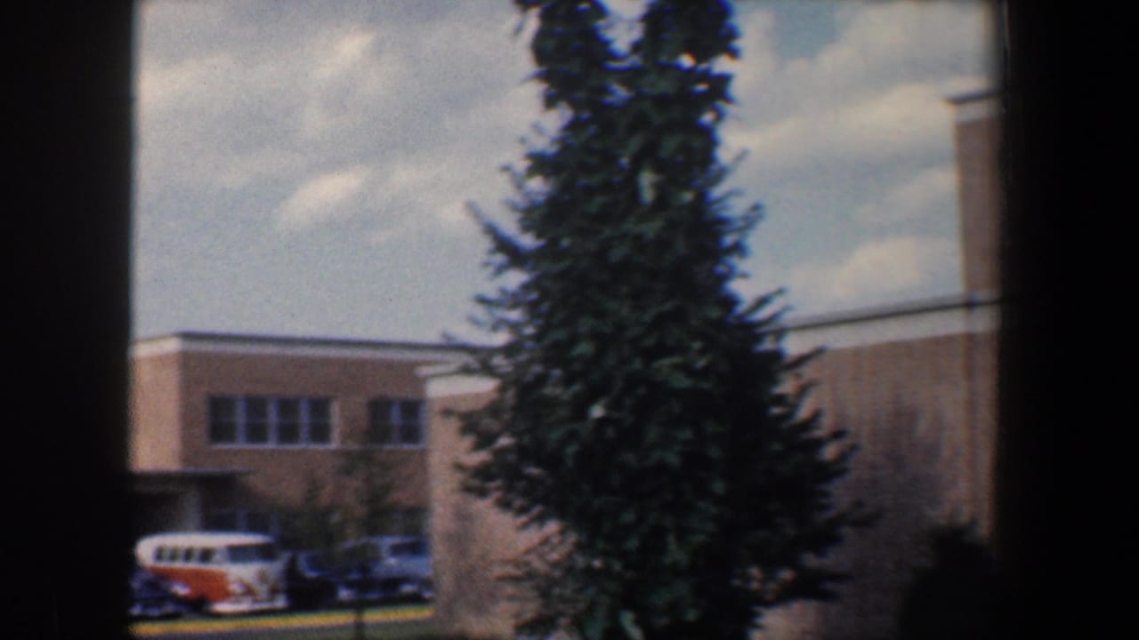 Tan brick building behind evergreen trees with parked cars and a small orange-and-white van.
