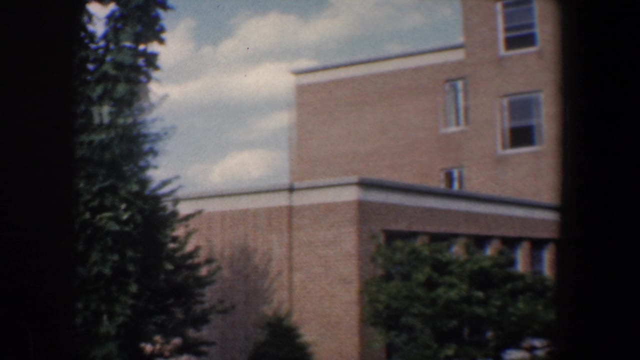 1960 Baltimore building exterior with tan brick walls, white-trimmed windows, evergreen trees, and visible 8mm gate shadows.