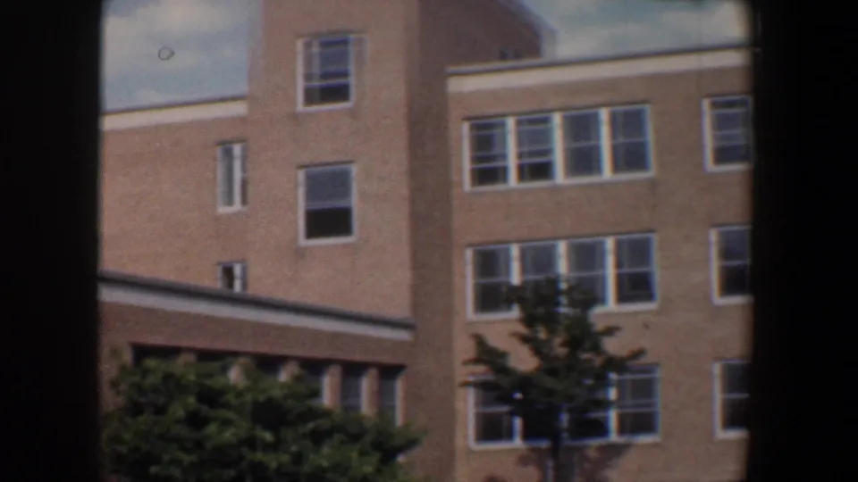 1960 Baltimore exterior showing a tan brick facade with rows of white-framed windows, landscaping, and 8mm frame shadows.