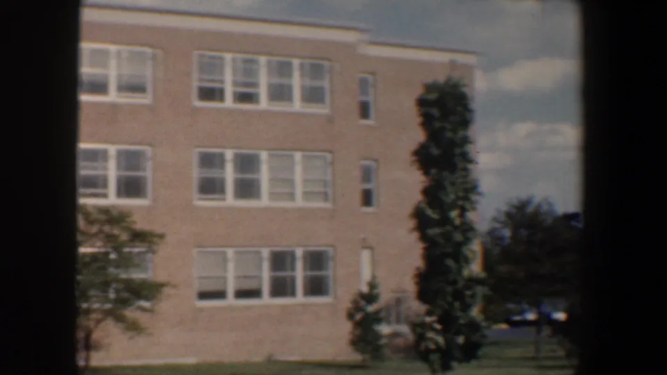 1960 Baltimore 8mm frame showing the side of a brick building, white-framed windows, trees, lawn, and partly cloudy sky.