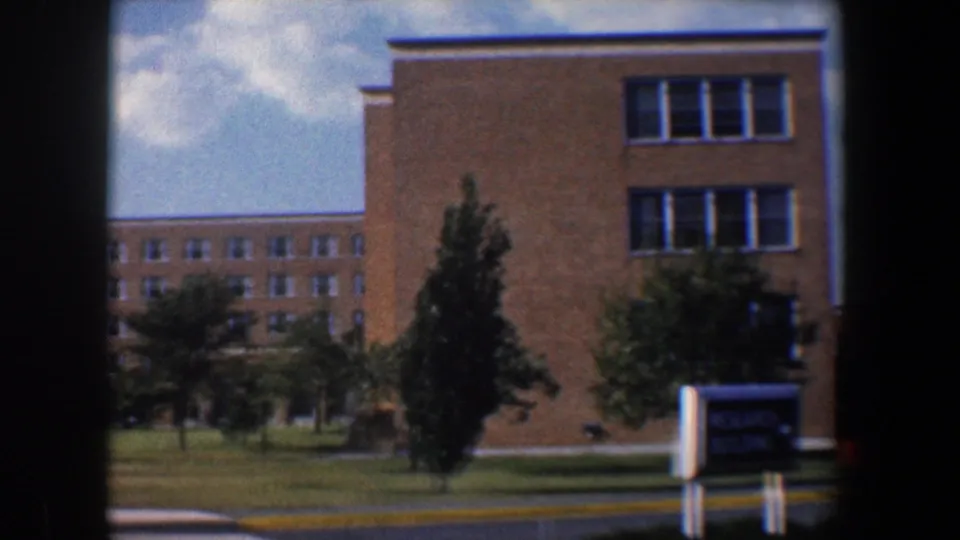 1960 Baltimore 8mm exterior showing brick buildings across a lawn with trees, blue sky, clouds, and an unreadable sign.