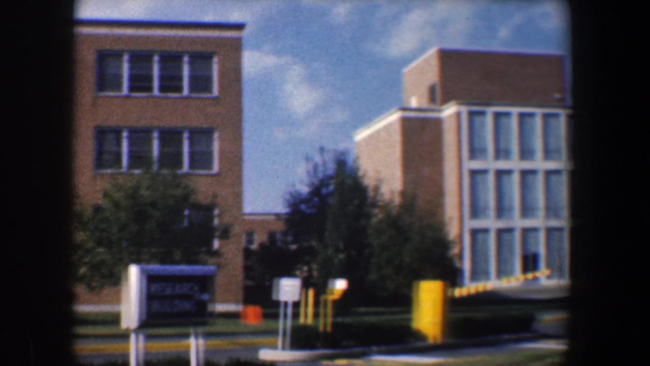 1960 Baltimore campus exterior showing a Research Building sign, tan brick facade, trees, and yellow parking-control posts.
