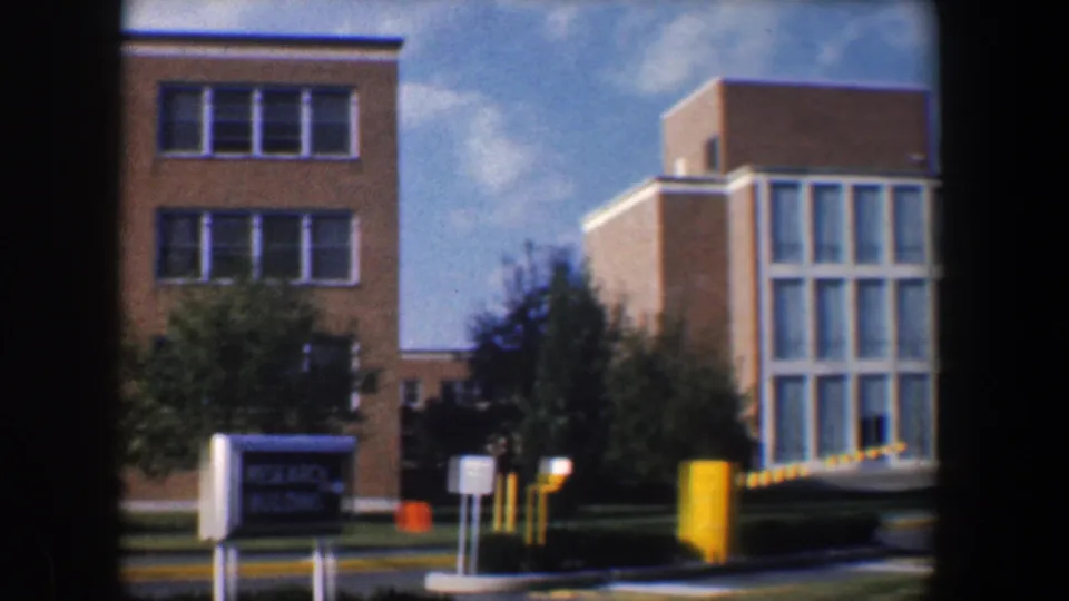1960 Baltimore campus exterior showing a Research Building sign, tan brick facade, trees, and yellow parking-control posts.