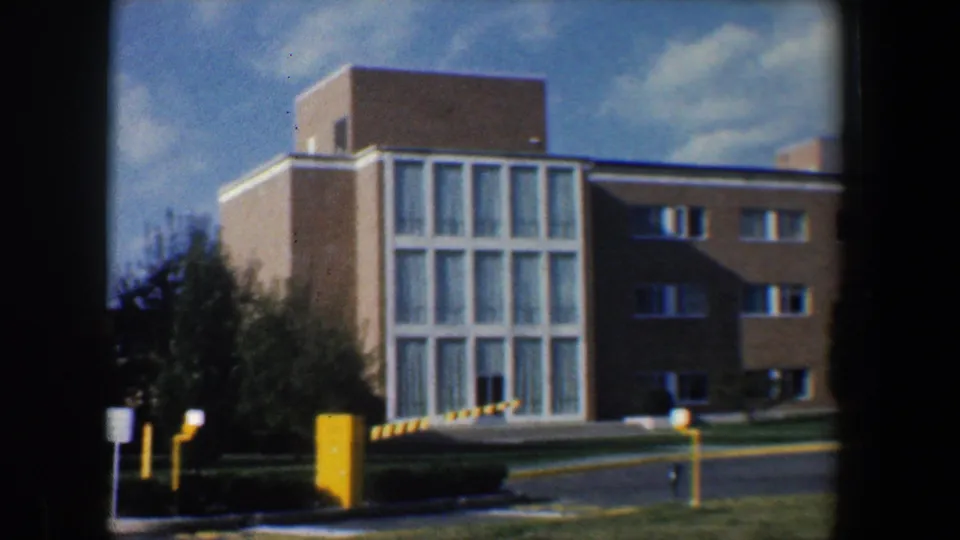 1960 Baltimore research-campus exterior with a tall glass window grid, tan brick walls, trees, and a yellow parking barrier.