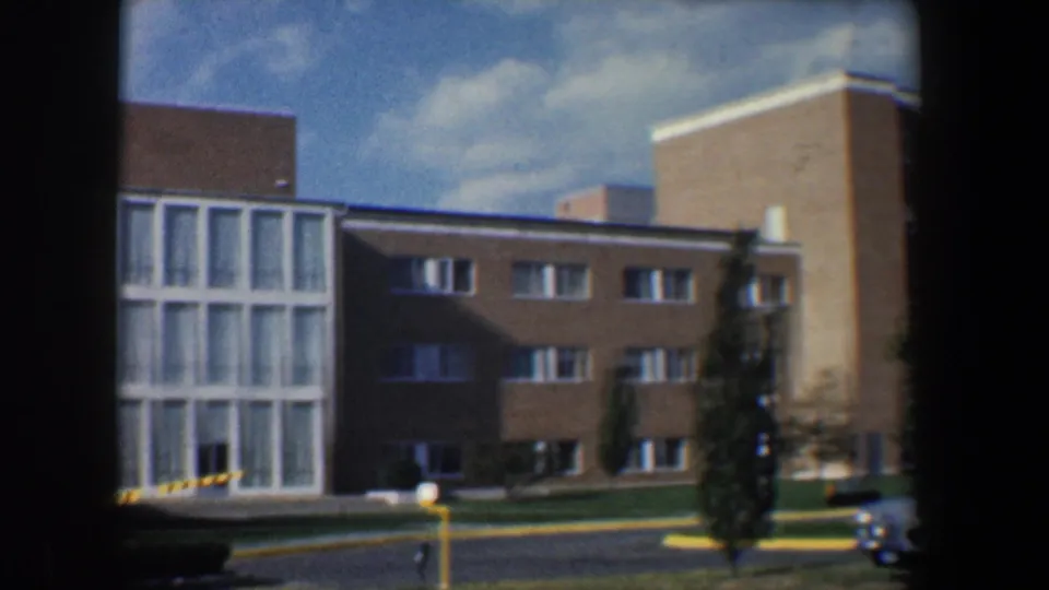 1960 Baltimore exterior view of a tan research-campus building with tall windows, roadway, lawn, and a small car at frame edge.