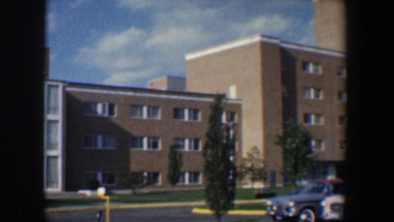 Brick multi-story apartment building with parked cars and small trees under a partly cloudy sky.