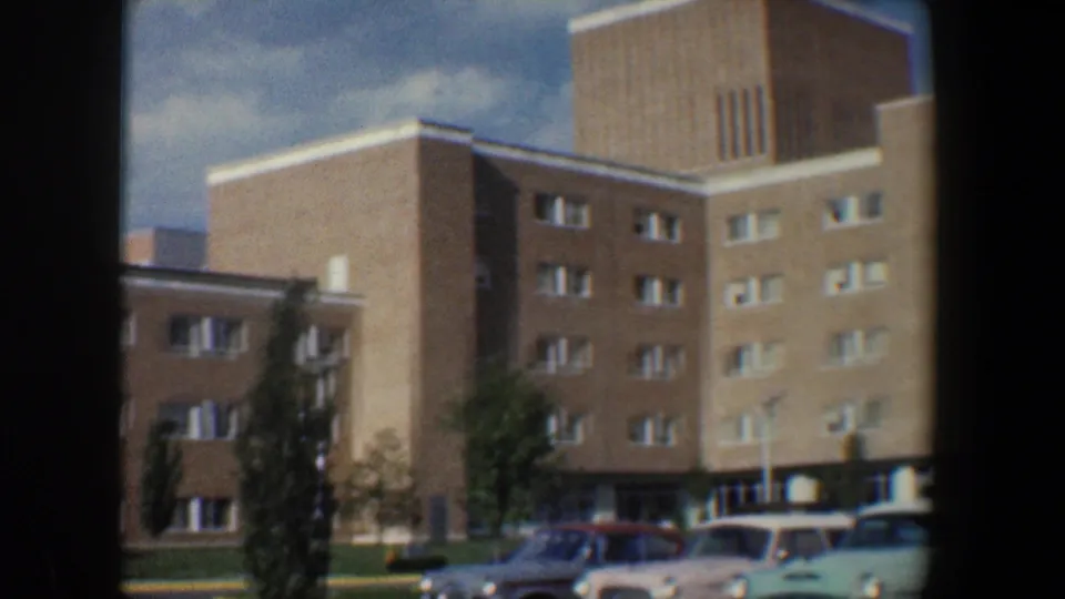 1960 Baltimore exterior with a multi-story brick apartment building, prominent tower, parked cars, curb, trees, and cloudy sky.
