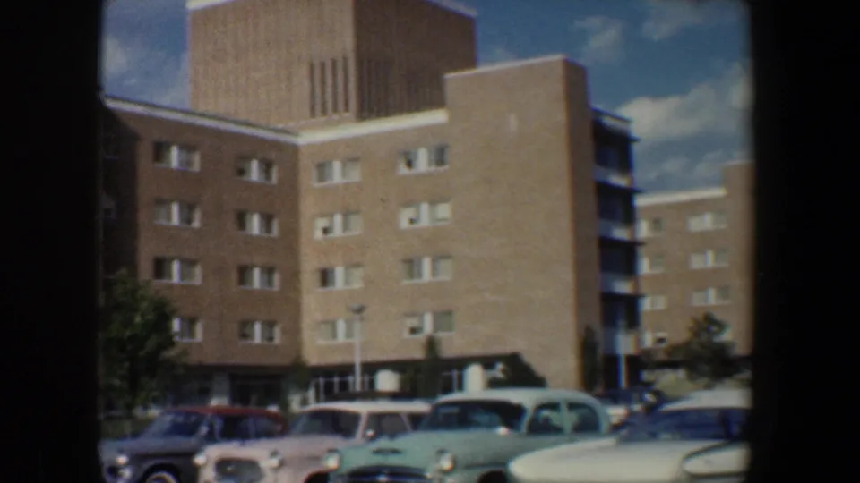 1960 Baltimore frame showing the apartment tower, side building, parked period vehicles, lawn, small trees, and overcast patches of sky.