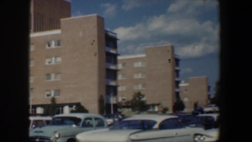 1960 Baltimore home-movie frame with brick apartment blocks, balconies or side openings, parked cars, trees, curb, and partly cloudy sky.