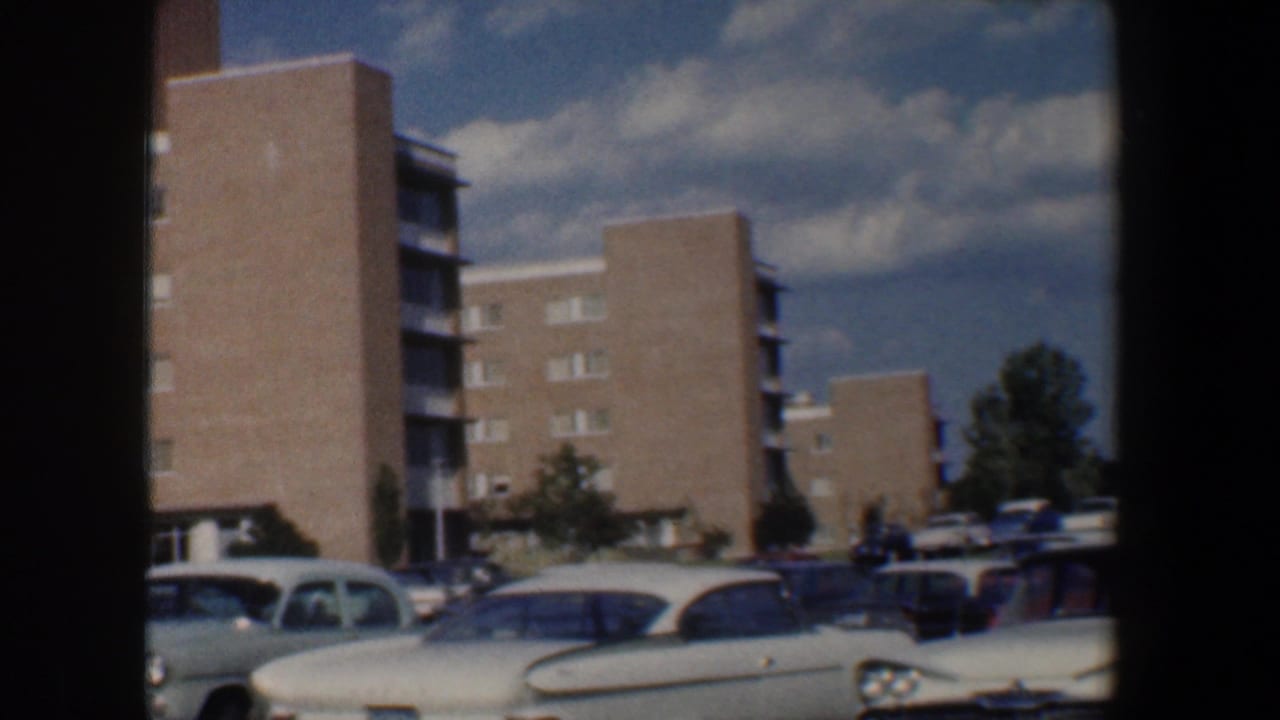 1960 Baltimore home-movie frame showing parked cars beside mid-century brick apartment buildings under a partly cloudy sky.