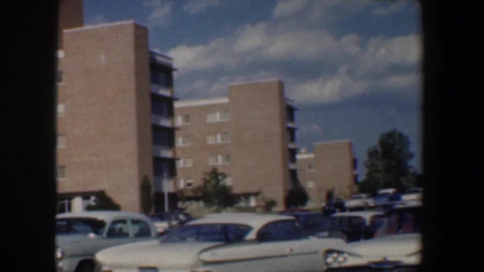 1960 Baltimore home-movie frame showing parked cars beside mid-century brick apartment buildings under a partly cloudy sky.