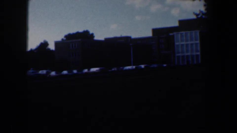 1960 Baltimore frame with apartment blocks, a line of parked cars, and a dark foreground from the panning 8mm shot.