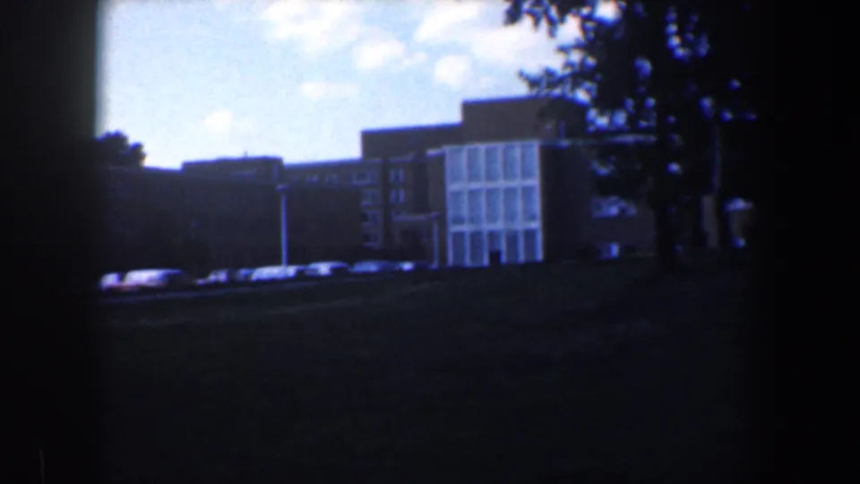 1960 Baltimore 8mm frame showing a glass-fronted apartment facade, parked cars, trees, and a dark grassy foreground.