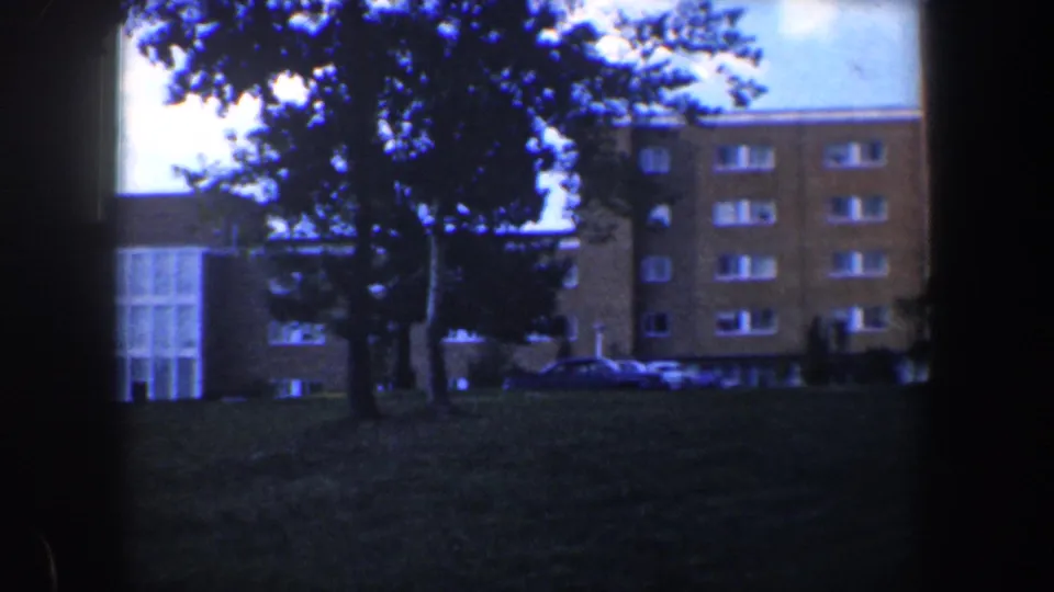 1960 Baltimore 8mm frame showing a brick apartment building, trees, lawn, and parked cars at the base.